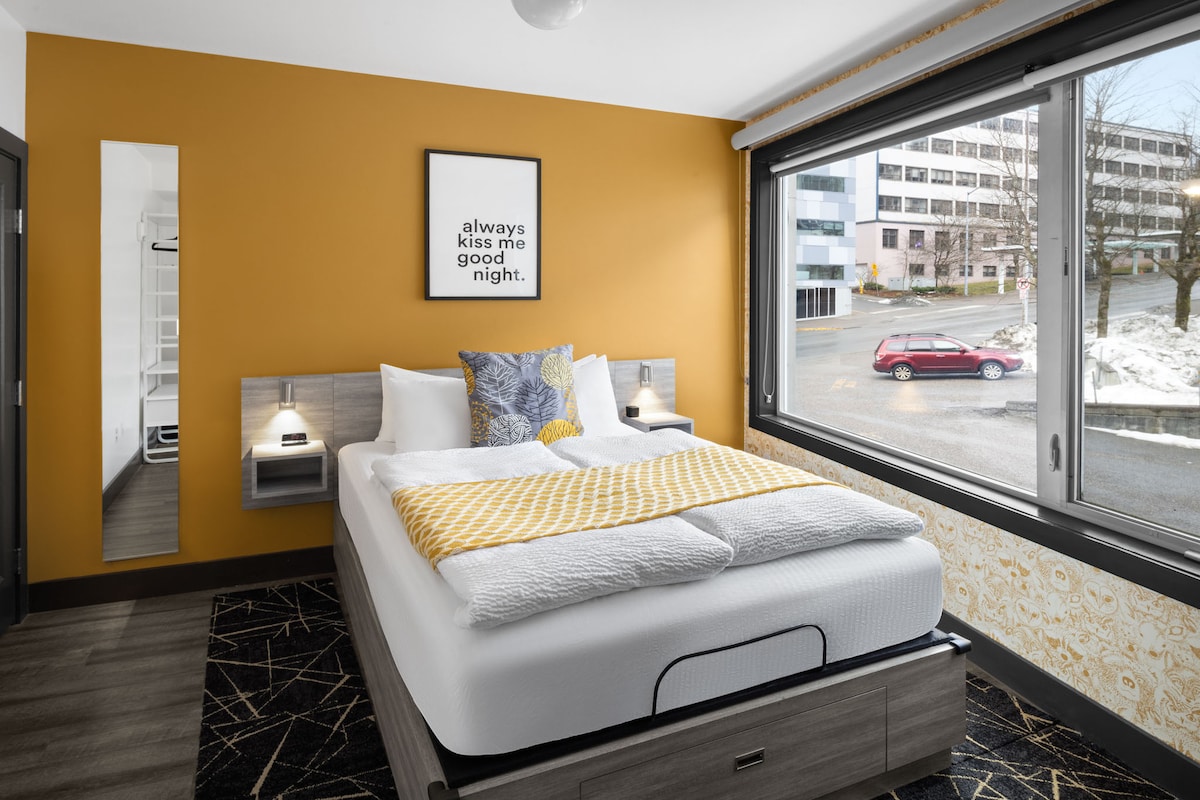 The room features a cozy queen bed with European-style bedding, adorned with a yellow accent blanket. Large windows provide views of the neighborhood, while a warm orange wall adds a pop of color. Two bedside lamps and a simple wall art piece contribute to the welcoming ambiance.