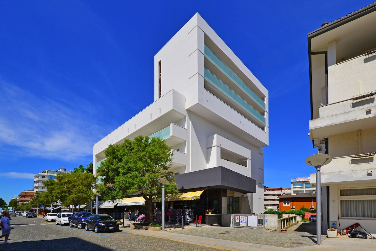 A modern building with a striking white facade is depicted, featuring multiple balconies and large windows. The structure stands at five stories and is surrounded by greenery and parked cars, indicating a lively urban environment close to the city center.