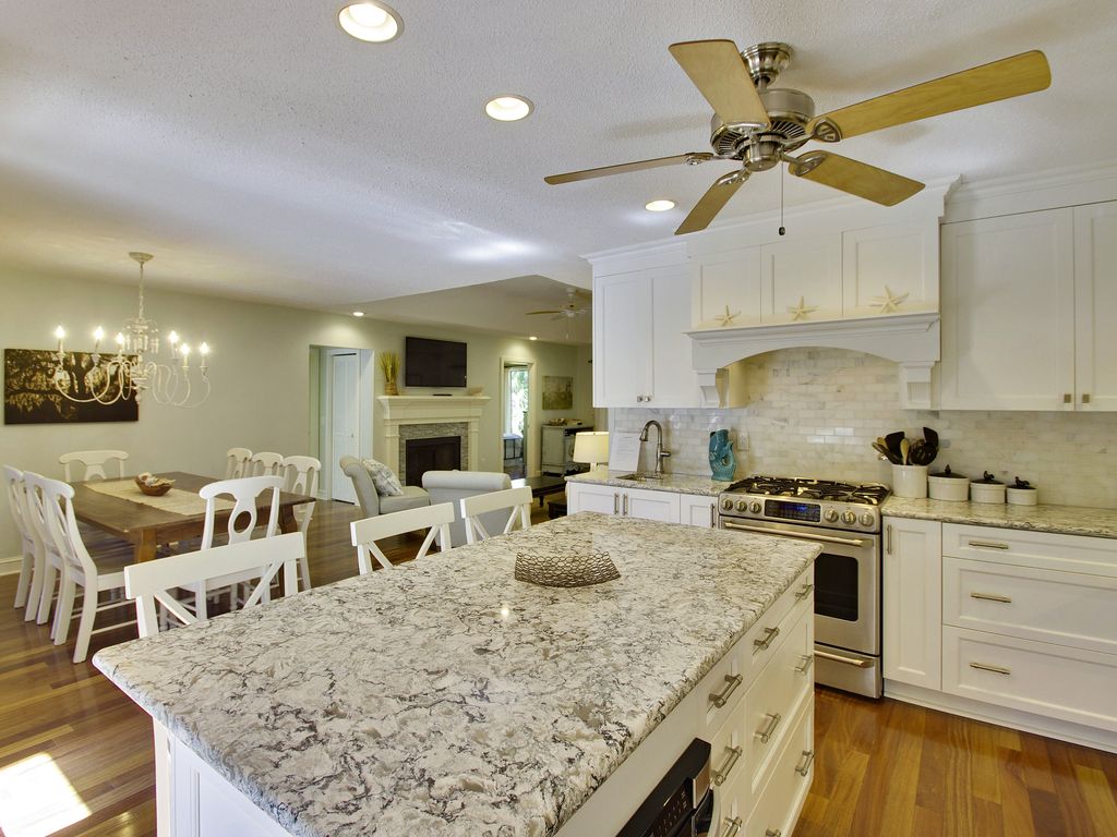 A spacious kitchen features a large granite island, accompanied by modern appliances including a gas range. The open layout connects to a dining area with a long table that seats up to 8. Soft lighting emanates from ceiling fixtures, enhancing the inviting atmosphere.