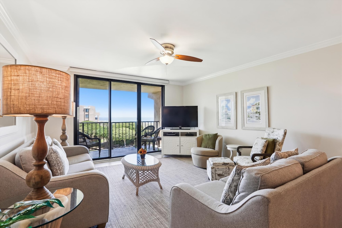 A spacious living area features comfortable seating arrangements, including two armchairs and a sofa. A flat-screen TV is mounted against the wall, with natural light streaming through large sliding glass doors that lead to a private balcony overlooking the ocean.