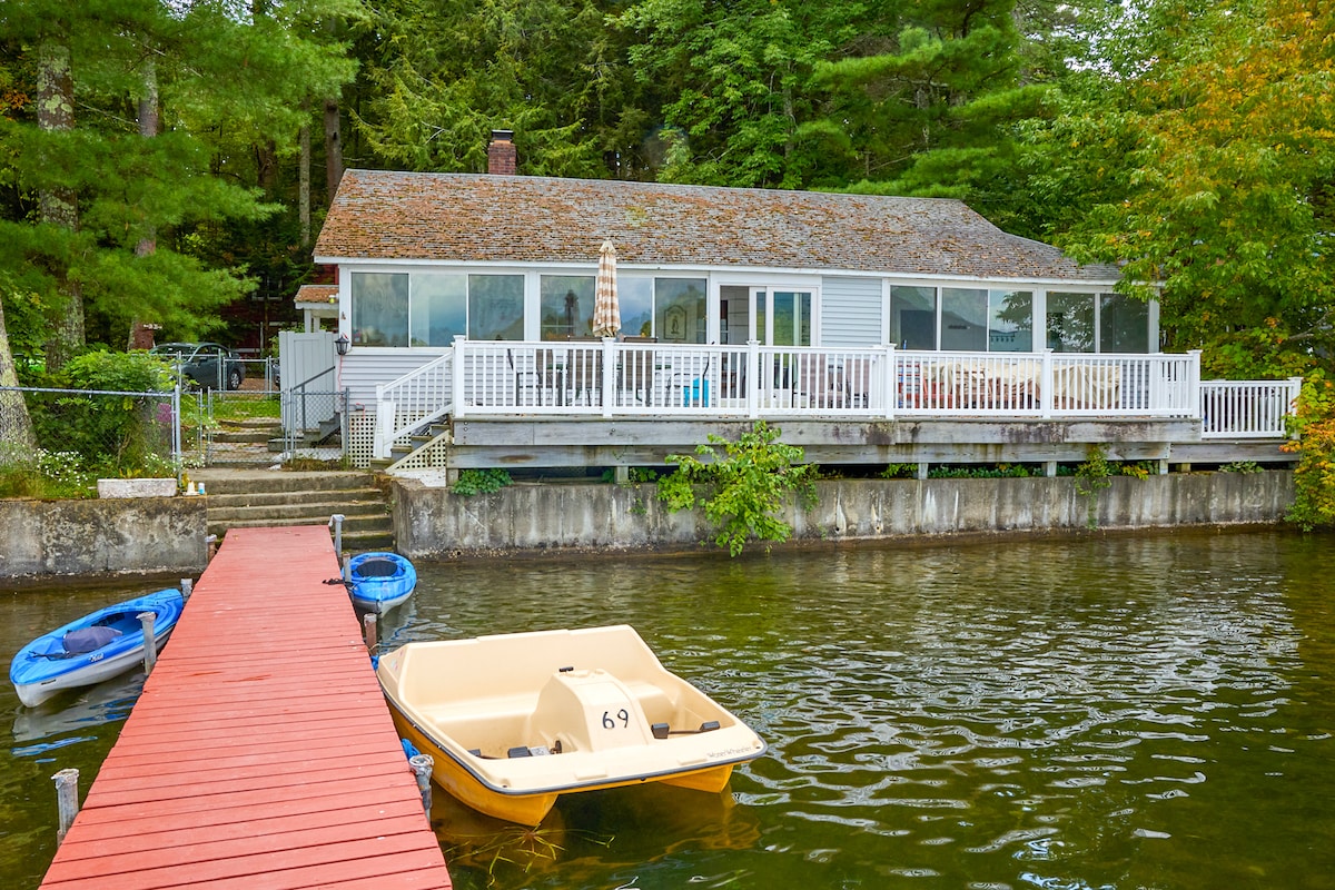 The lakefront property is depicted featuring a spacious deck with a safety gate, perfect for outdoor activities. A red dock extends into the water, accommodating a pedal boat and two kayaks. Lush greenery surrounds the area, enhancing the serene lakeside environment.