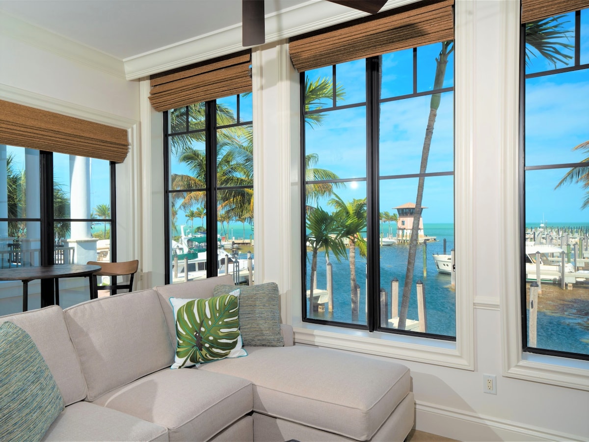 A spacious living area is enhanced by large windows, offering views of palm trees and the marina. The light-colored sectional sofa features decorative pillows, and a small dining table is visible in the background, creating a comfortable and inviting atmosphere.
