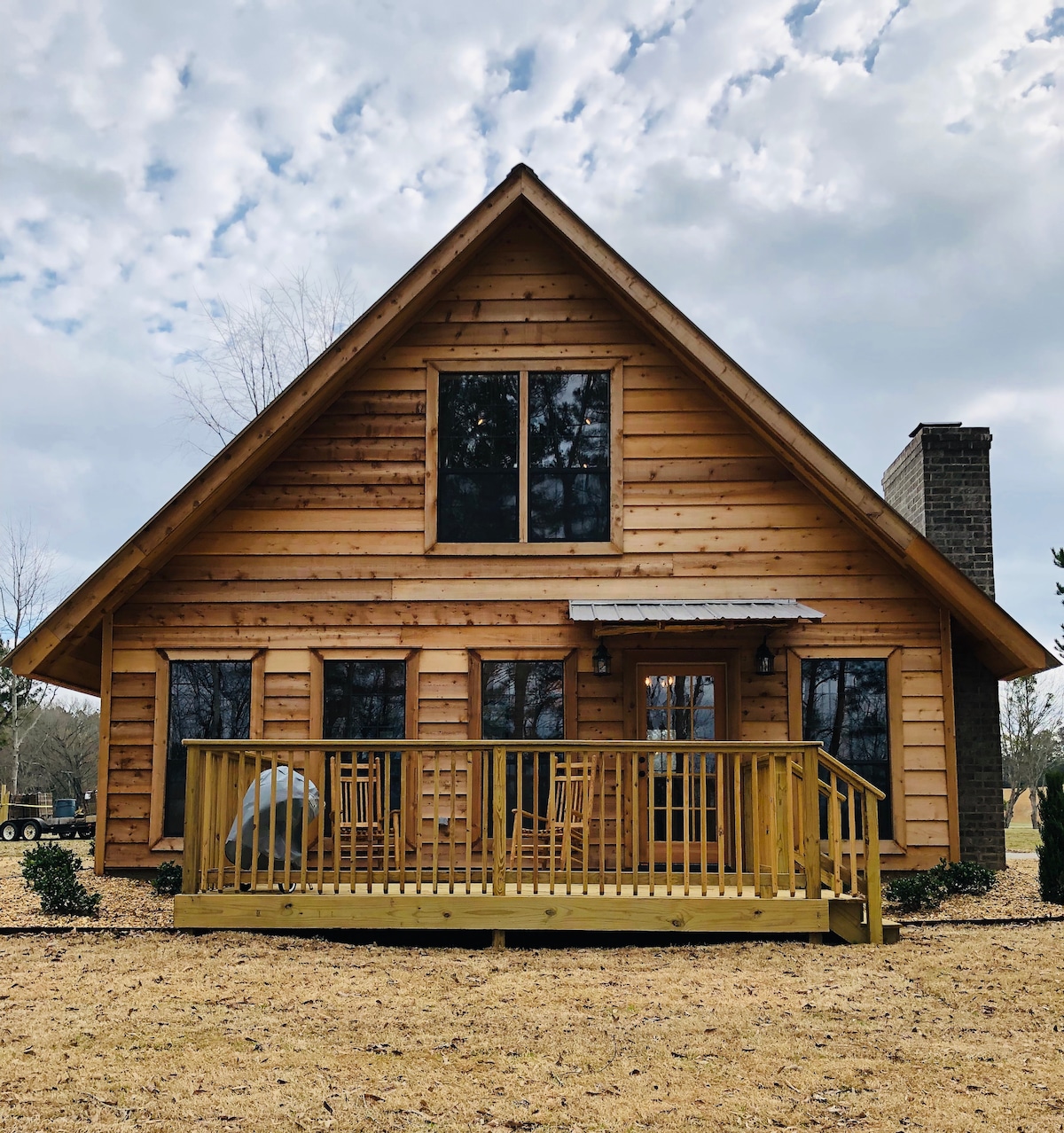 A rustic cabin with wooden siding features a steeply pitched roof and multiple windows. The front porch is adorned with wooden railings and seating. A stone chimney rises from the right side, framed by neatly landscaped areas, set against a cloudy sky.