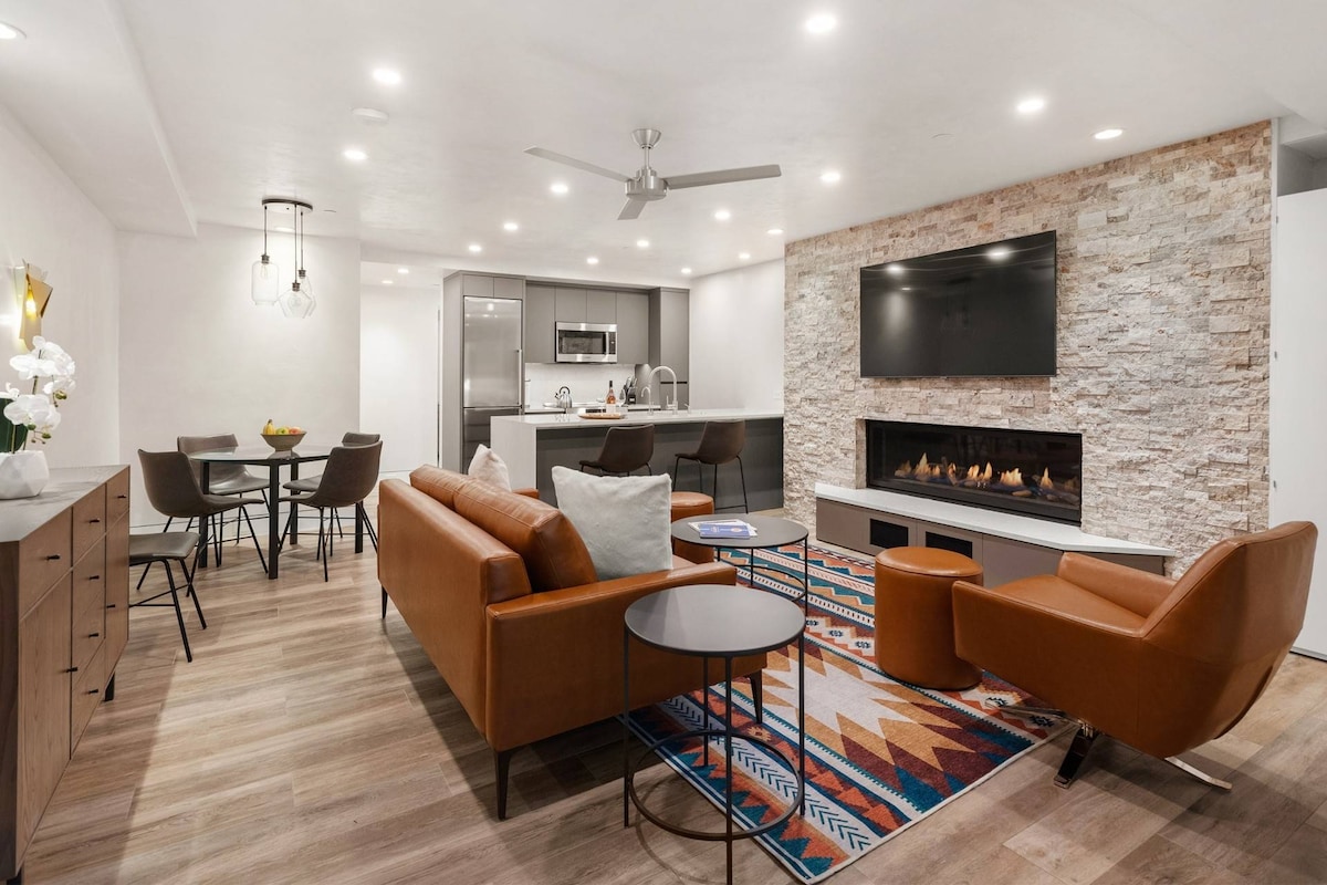 A modern living area is highlighted by a gas fireplace set within stone accents. Comfortable seating is provided by leather sofas, complemented by a patterned rug. A dining table for four and a well-equipped kitchen are also visible, with ample lighting enhancing the open space.