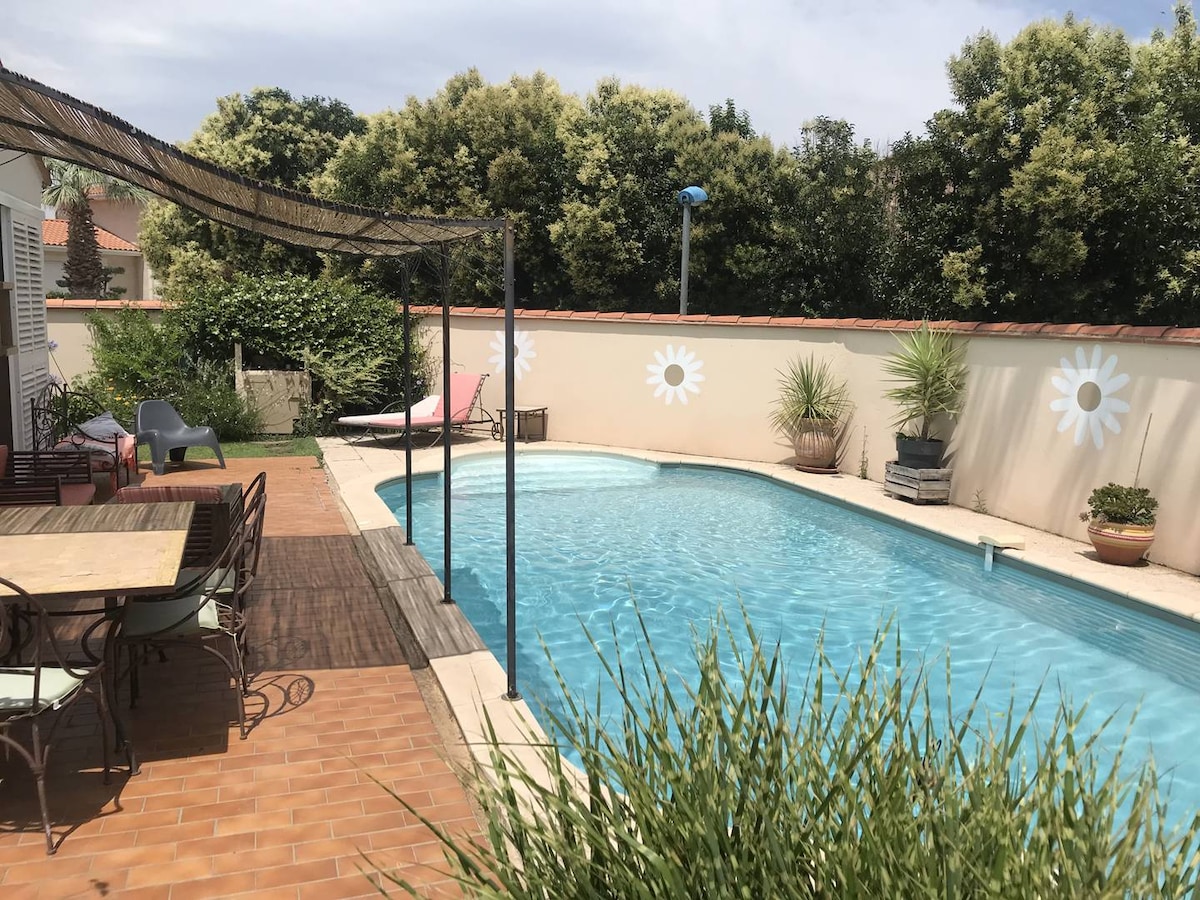 A private rectangular pool is surrounded by a wooden deck, featuring a shaded dining area with a wooden table and chairs. Lush greenery and potted plants enhance the space, while a sunbed and loungers are positioned nearby, providing a tranquil outdoor retreat.