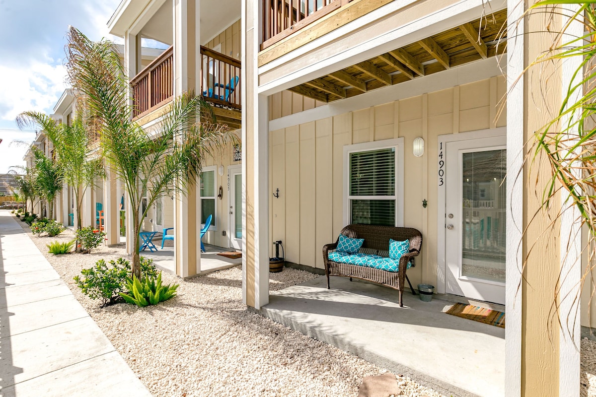 The exterior features a covered porch with a comfortable seating area adorned with blue cushions. Lush greenery, including palm plants, frames the entrance, while a gravel pathway leads to the door. The townhouse has a welcoming, well-maintained appearance.