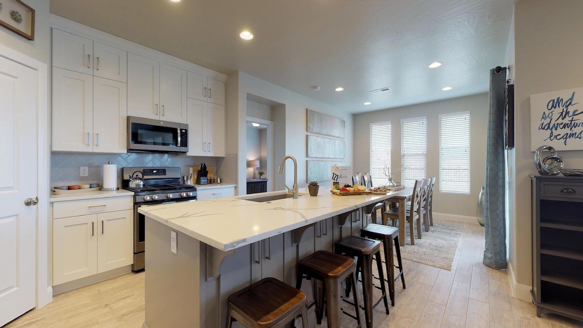 A modern kitchen is featured with white cabinetry and stainless steel appliances. A large island with bar seating offers ample space for dining. A dining area with a long table and multiple chairs is visible, along with large windows allowing natural light to brighten the space.