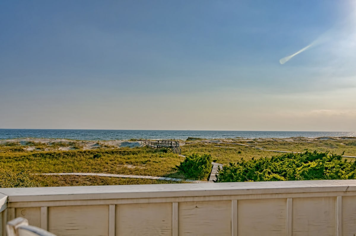 A sweeping view of the Atlantic Ocean stretches across the horizon, with gentle waves lapping at sandy shores. Lush greenery frames the foreground, leading toward a sandy path that guides the eye toward the beach. The sky is illuminated with soft colors from the setting sun.