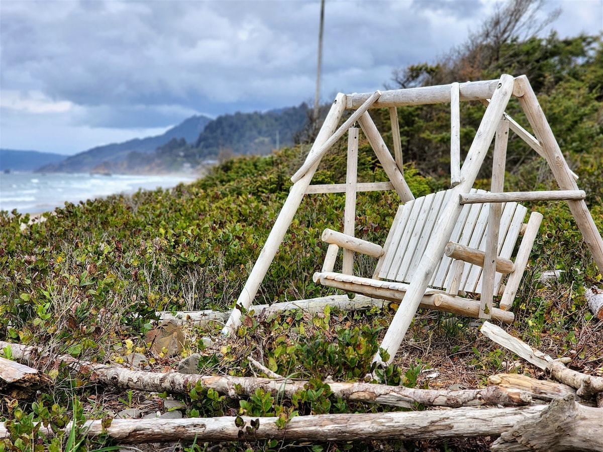 A rustic wooden swing is positioned amid coastal greenery, offering views of the ocean in the distance. Weathered logs surround the swing, enhancing the natural setting. Dark clouds loom overhead, hinting at the nearby shore and the tranquil ambiance of the area.