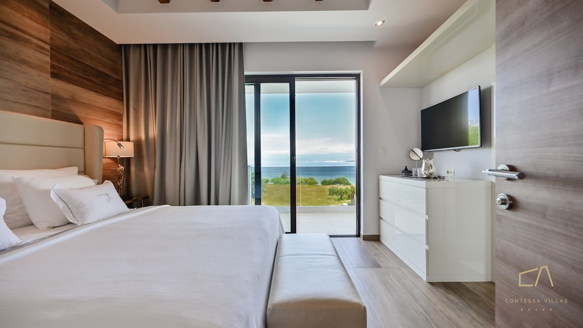 A serene bedroom features a spacious bed framed by minimalist wood detailing. Large glass doors offer panoramic sea views and access to an outdoor area. A sleek television and ample storage space are integrated into the design, enhancing the room's modern aesthetic.