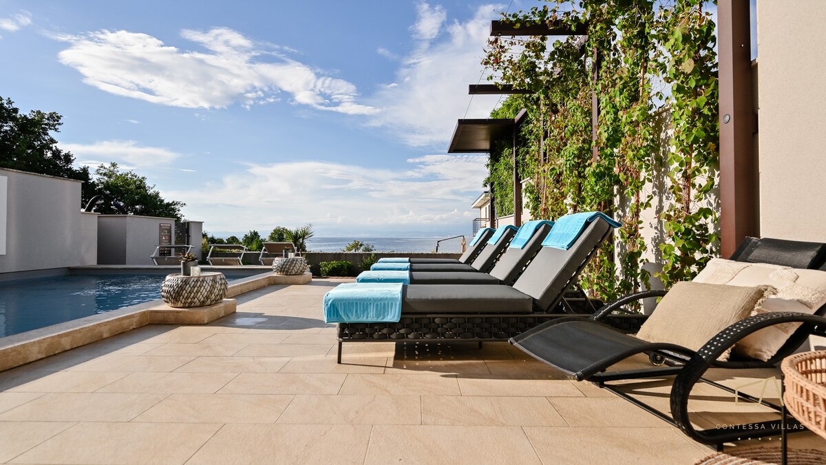 A spacious outdoor area is presented with comfortable lounge chairs adorned with blue cushions, positioned near a private swimming pool. Lush greenery frames the space, creating a serene atmosphere with views of the sea in the background under a bright sky.