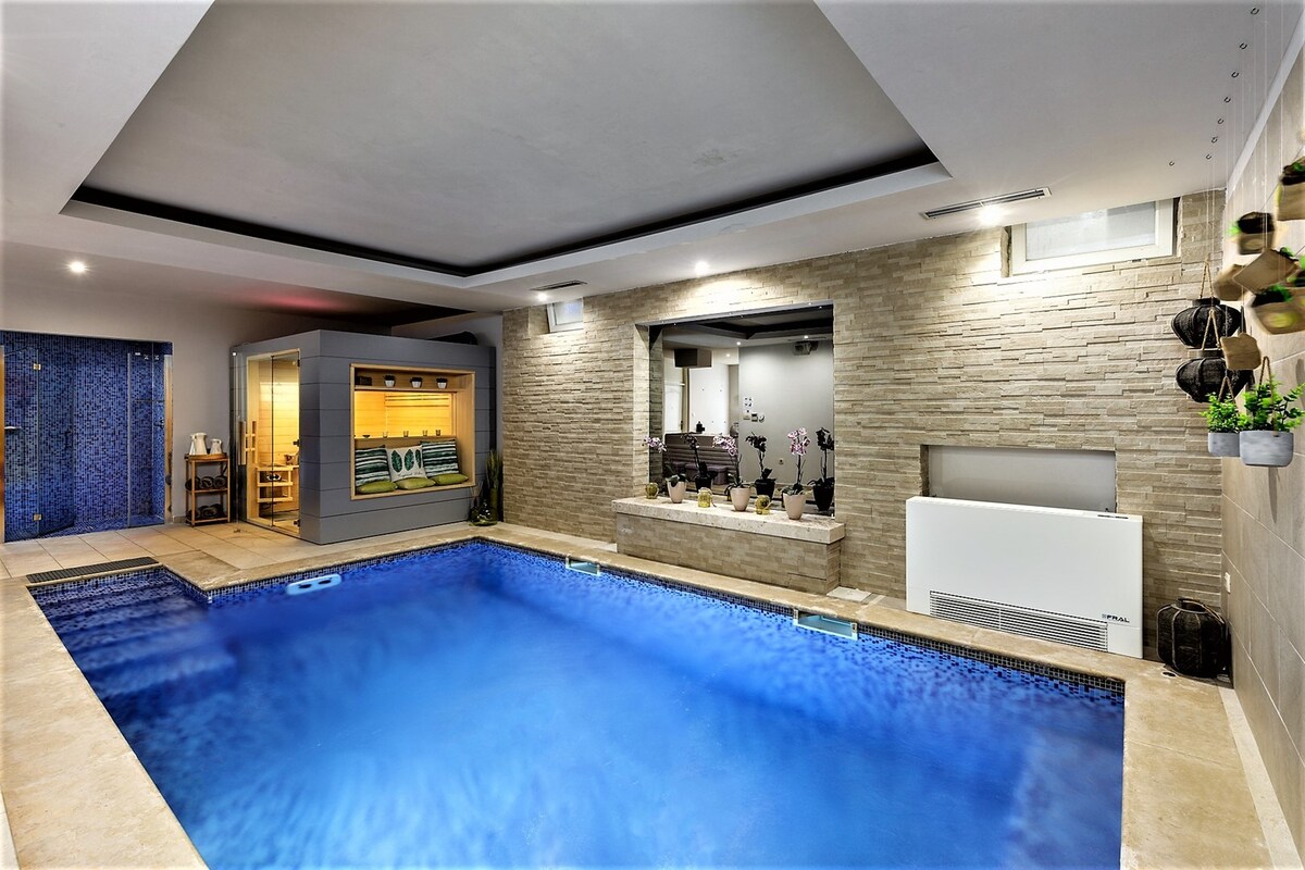 An indoor pool area features a clear blue swimming pool, surrounded by light-colored stone walls. Natural light filters in through a large window near the far end, where a sauna and seating area are also present, inviting relaxation and tranquility.