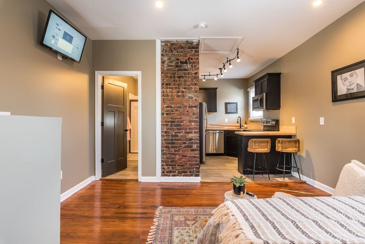 The well-appointed studio features a blend of modern amenities and rustic charm. A textured brick wall provides visual interest, while a kitchenette with stainless steel appliances is visible. Natural light enhances the inviting atmosphere, complemented by wood flooring and tasteful decorations.