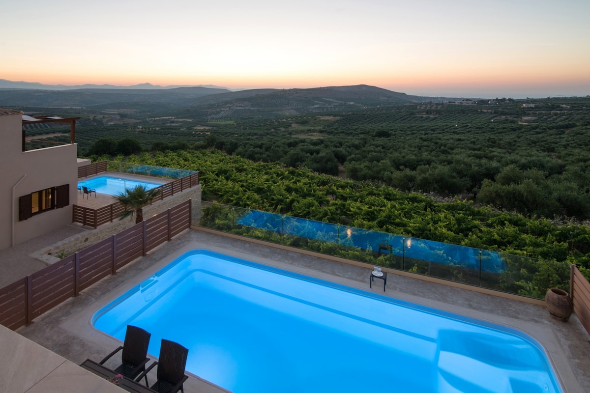 A clear view captures two inviting heated pools nestled beside lush vineyards, with rolling hills in the background. The soft glow of dusk highlights the tranquil landscape, enhancing the serene setting. Comfortable chairs are positioned near the water’s edge, creating an ideal relaxation spot.