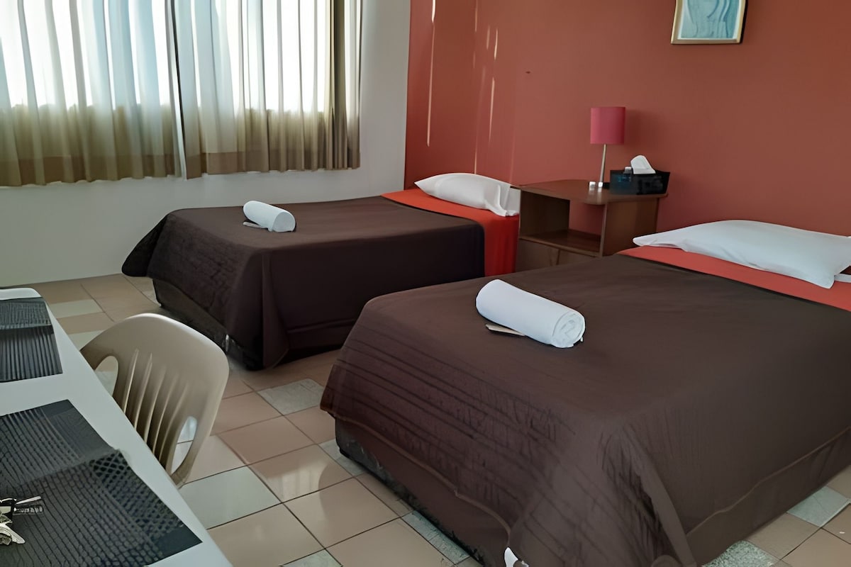 The room features two spacious beds, each draped with dark bedding. A small table with a lamp and a tissue box is positioned beside one bed. Light filters through large, sheer curtains, creating a bright atmosphere in the space.