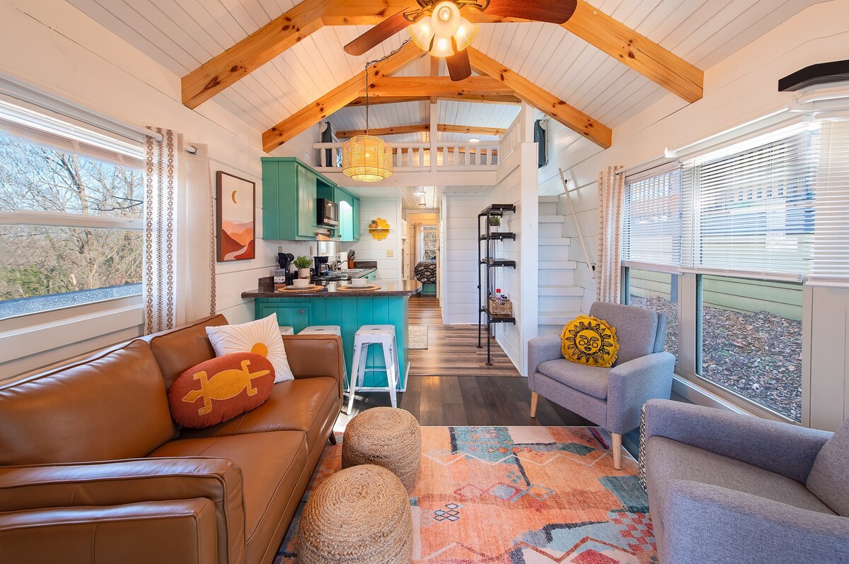 The living area offers a blend of comfort and style, featuring a brown leather sofa paired with a light gray armchair. Natural light illuminates the room through large windows, highlighting the vaulted ceiling adorned with wooden beams. A colorful area rug adds a touch of warmth.