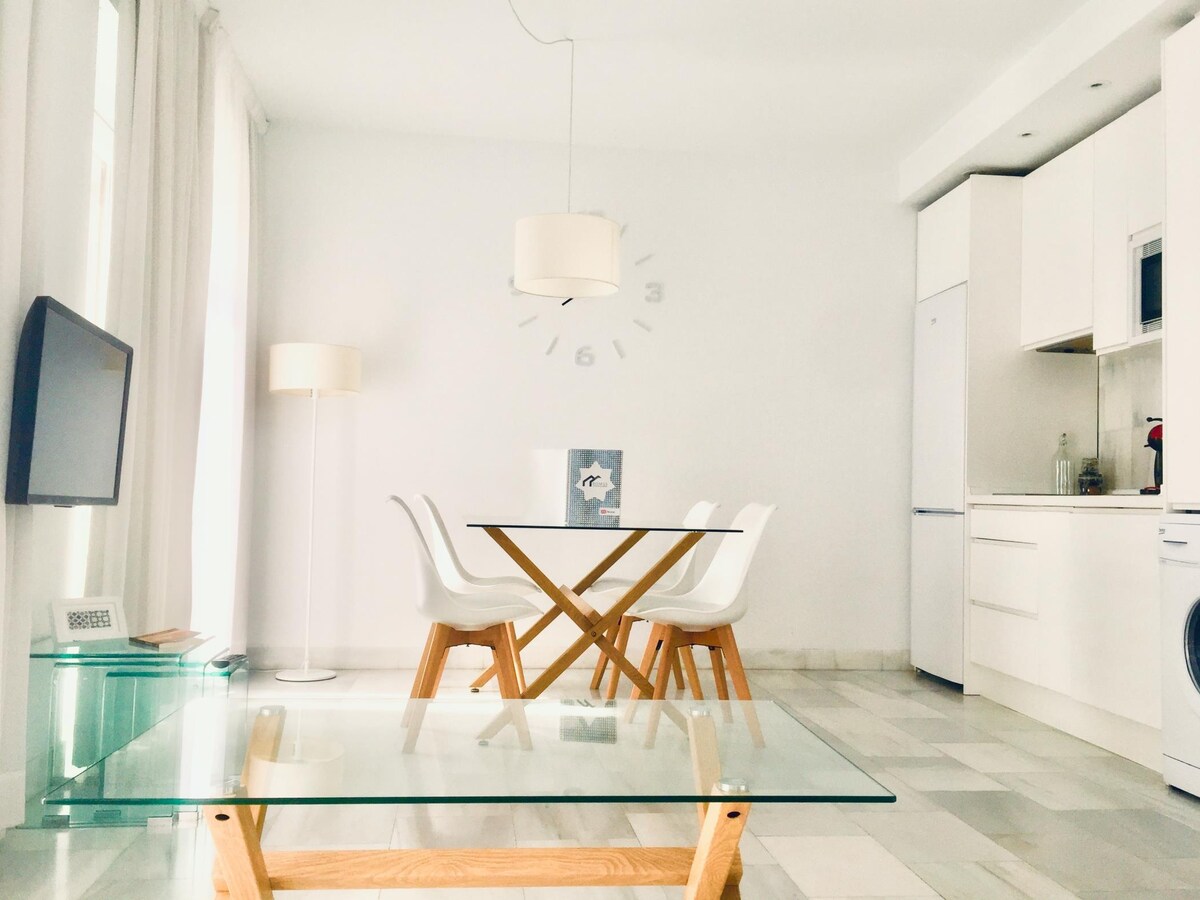 A modern living space features a transparent coffee table with wooden legs, complemented by a wooden dining table surrounded by four white chairs. A sleek television is mounted on the wall, while natural light fills the room through large windows adorned with sheer white curtains.