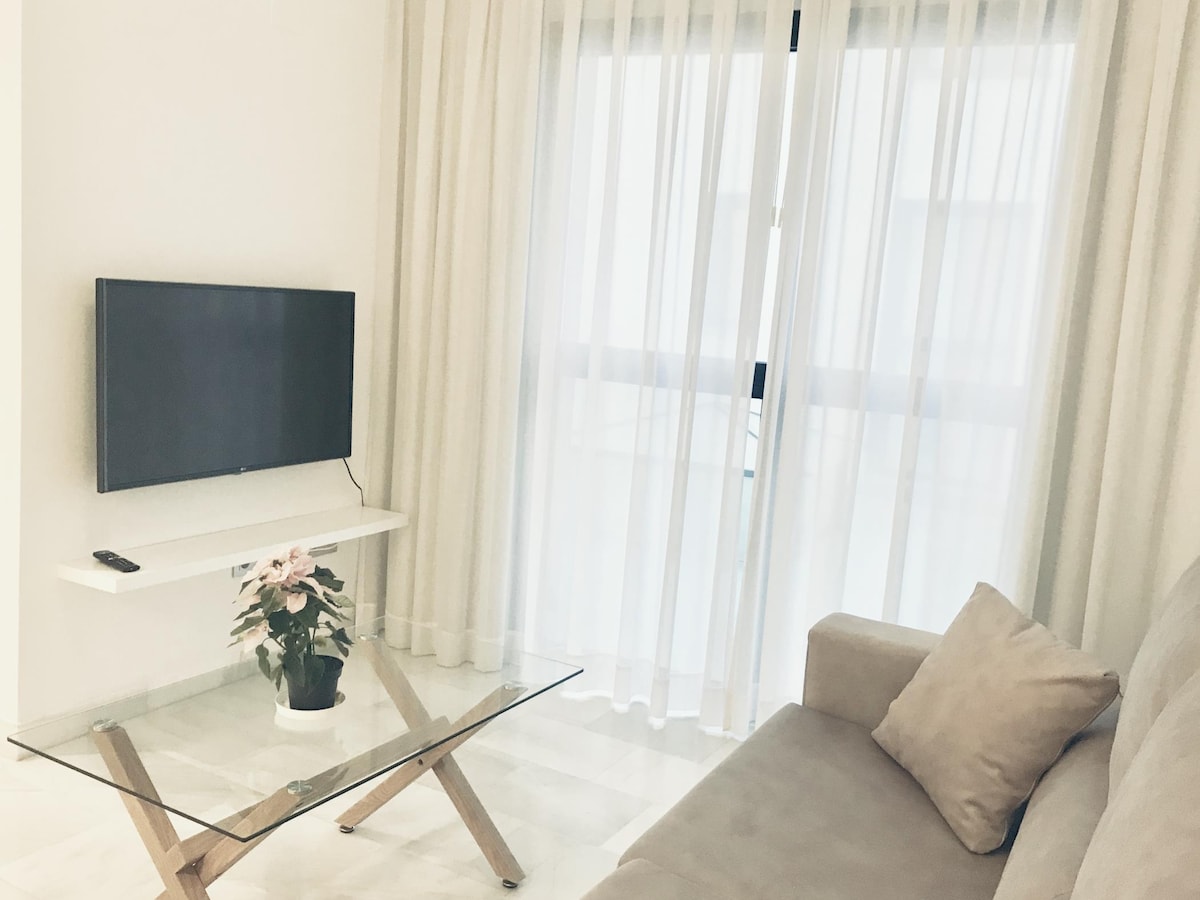 A bright living area features a comfortable sofa and a glass coffee table. A flat-screen TV is mounted on the wall, and sheer curtains allow natural light to gently filter in, creating a serene atmosphere. A small potted plant adds a touch of greenery to the space.