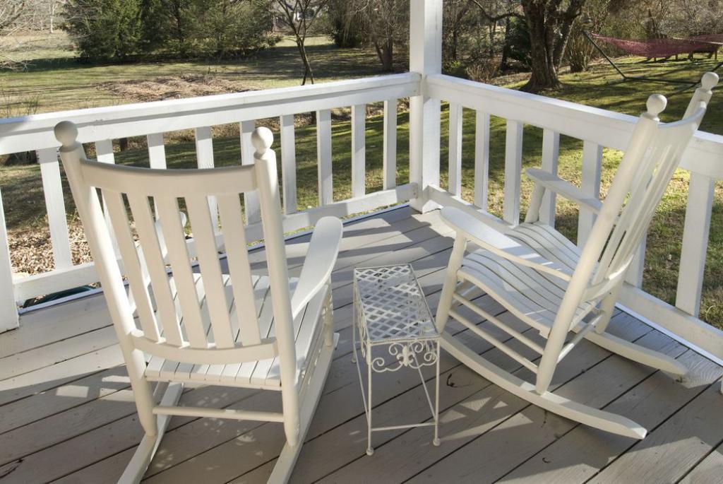 Two white rocking chairs and a small iron side table are positioned on a covered porch, overlooking a serene natural landscape. The porch railing and floor showcase a clean, light-colored wood finish, creating a peaceful outdoor space.