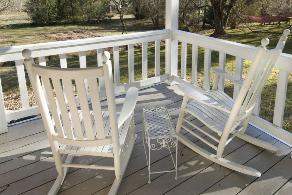 A peaceful porch setting features two white rocking chairs positioned around a small iron side table. The area overlooks a grassy landscape dotted with trees, inviting relaxation and moments of tranquility.