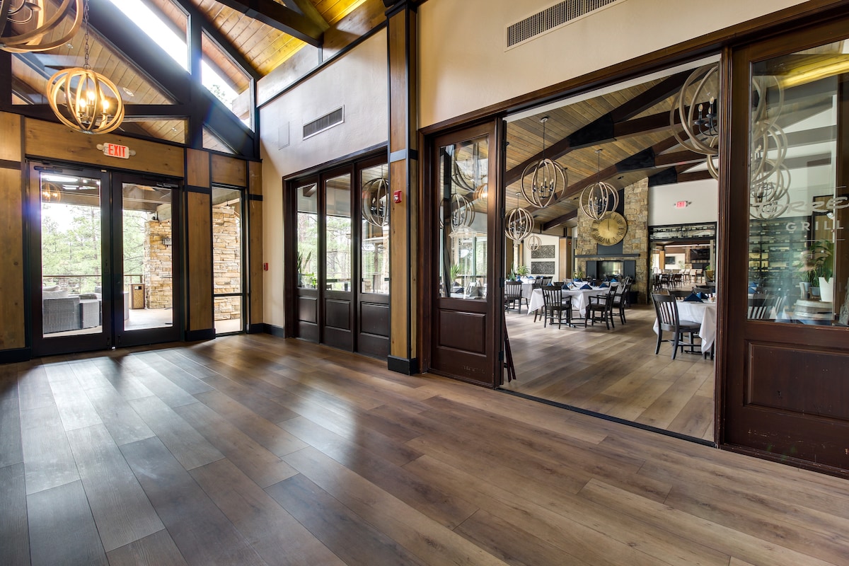 An open dining area is featured with large windows allowing natural light to fill the space. Modern light fixtures hang from the ceiling, complementing the warm wood tones of the flooring and beams. A glimpse of an outdoor area is visible through the doors.