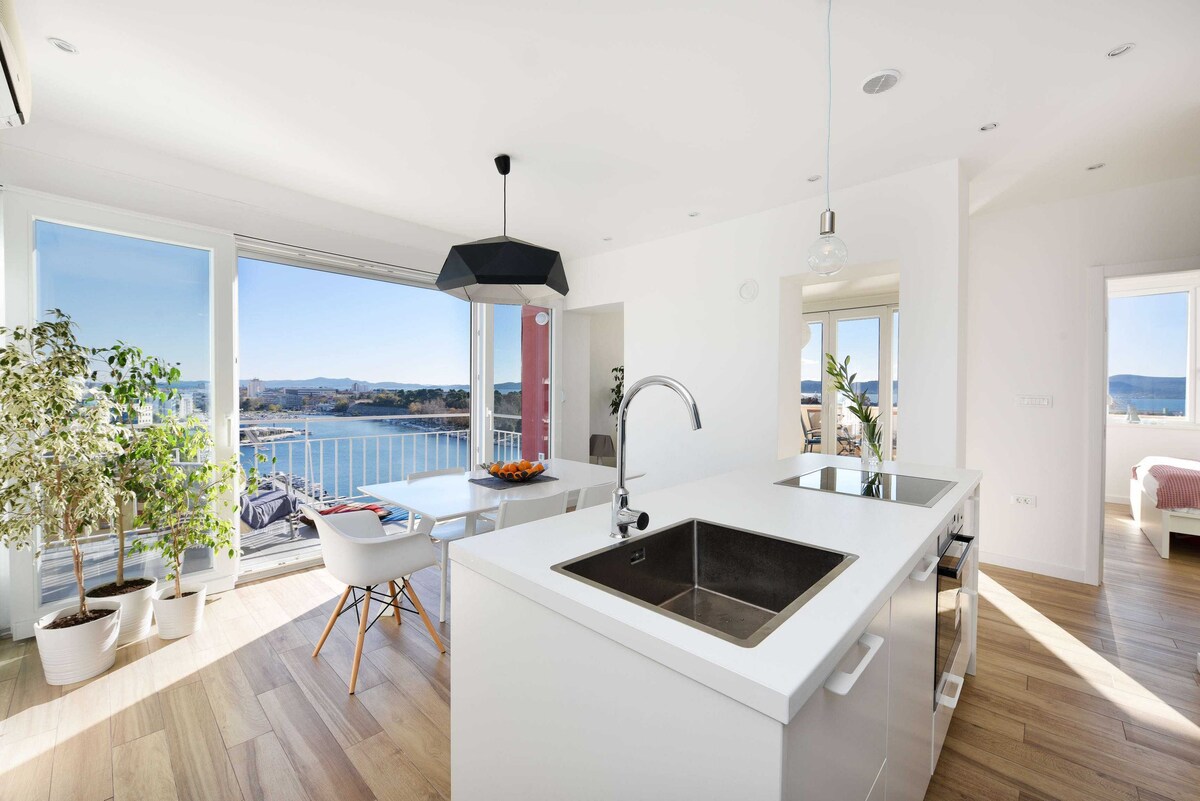 A modern kitchen area is visible, featuring a sleek island with a double sink and stylish fixtures. Large windows provide ample natural light and offer expansive views of the sea and surrounding landscape. A dining table with chairs is positioned nearby, enhancing the open layout.