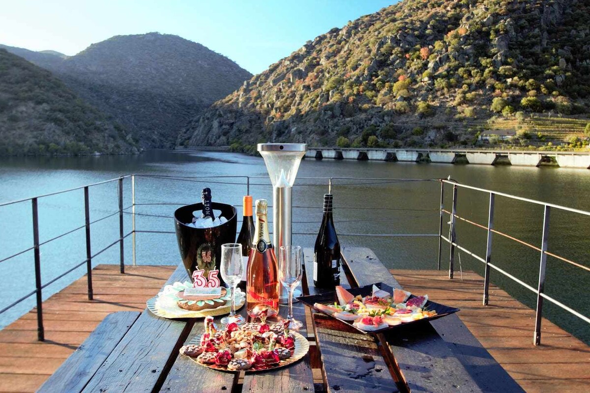 A dining table is set on a wooden dock, featuring an arrangement of various drinks, including a bottle in an ice bucket, and a selection of snacks. The serene waters reflect the surrounding mountains, creating a tranquil atmosphere.