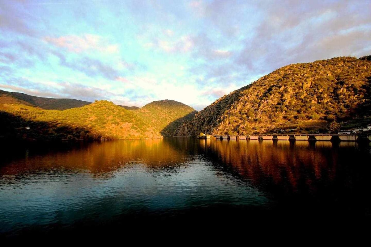 A tranquil river scene is presented, featuring calm waters reflecting the surrounding hills. Lush greenery and rocky slopes frame the shoreline, while a gentle gradient of clouds adds texture to the sky. The sunlight casts warm hues on the landscape, enhancing the peaceful ambiance.