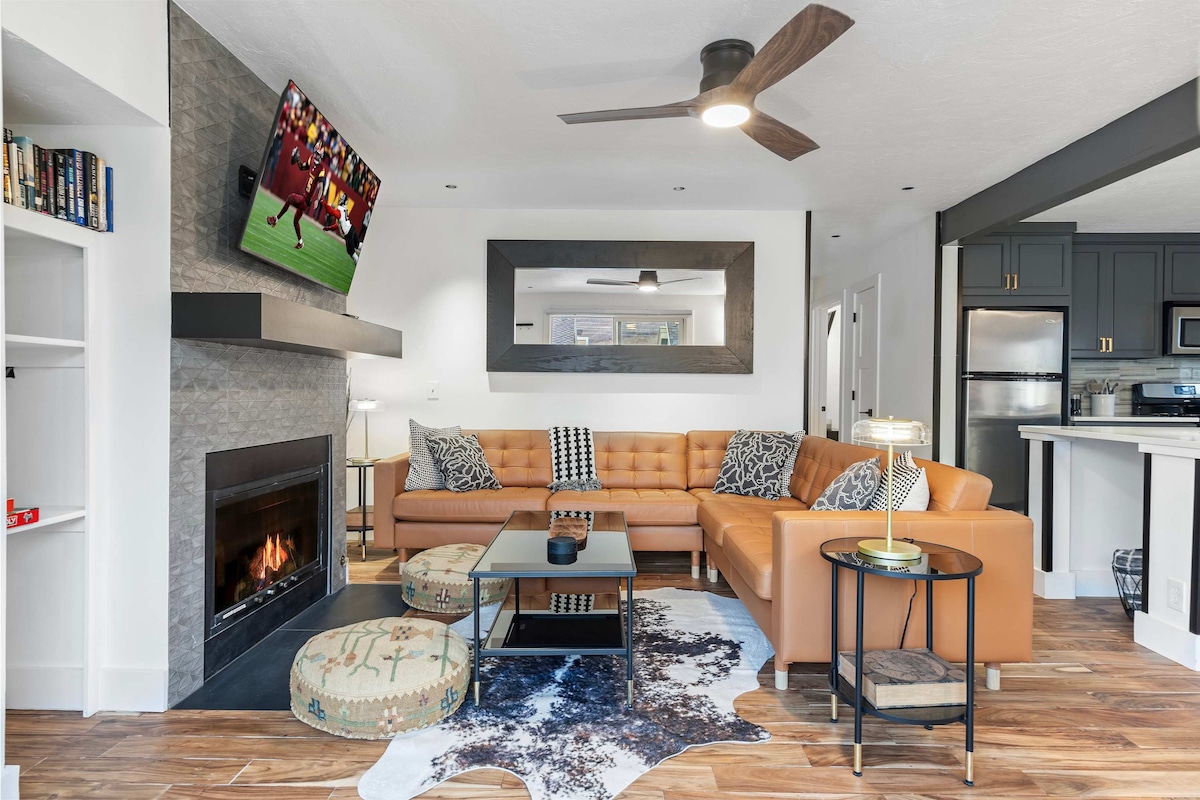 A modern living area is presented with a brown leather sectional couch and a glass coffee table. A television is mounted above a stone fireplace, with light-colored walls and natural wood flooring enhancing the space. A ceiling fan adds to the fresh atmosphere.