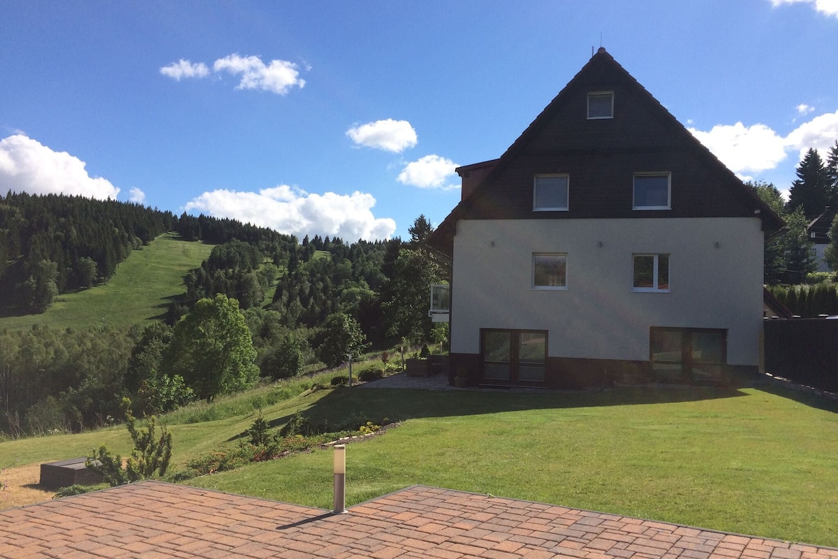 A spacious two-story villa is set against a backdrop of lush green hills and fields. The structure features large windows, allowing natural light to fill the rooms. Nearby ski slopes are visible, highlighting the outdoor recreational opportunities in the area.