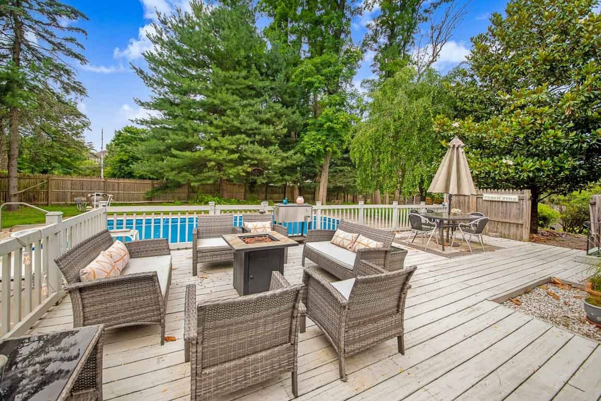 A spacious deck features comfortable seating arrangements with wicker chairs surrounding a central fire pit. The saltwater pool is visible in the background, framed by verdant trees and a fenced yard, providing a serene outdoor space for relaxation.