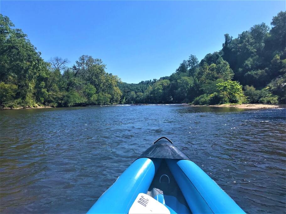 A view from a kayak on the Tuckasegee River is displayed, showcasing calm water surrounded by lush green trees on either bank. The bright blue sky reflects the serene natural landscape, inviting exploration and relaxation in the peaceful surroundings.
