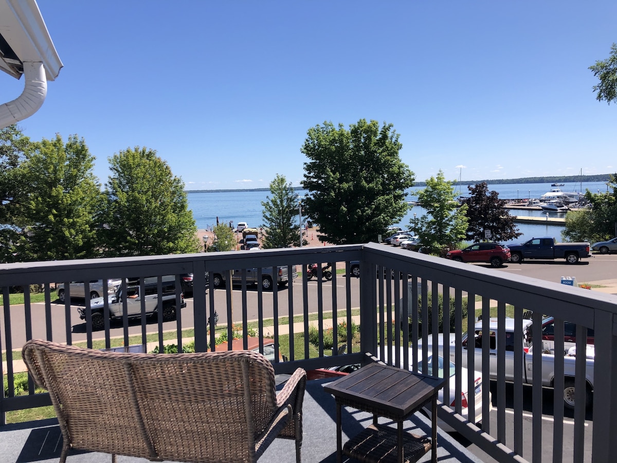 A private balcony features a wicker chair and small table, overlooking the scenic view of Lake Superior. Lush trees and a marina are visible in the distance, while clear skies invite relaxation. The space is designed for enjoying the outdoors.