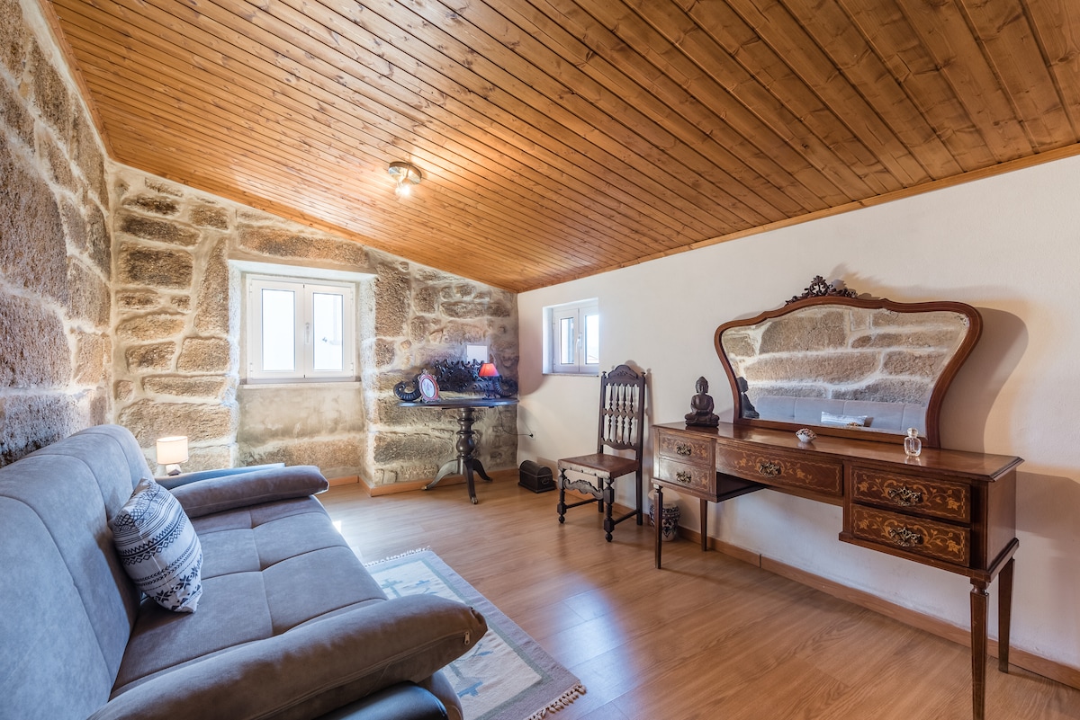 A cozy sitting area features natural stone walls and wooden ceiling beams. A sofa is positioned near two small windows that allow natural light. An antique wooden dresser and a vintage chair add charm, creating an inviting atmosphere for relaxation.