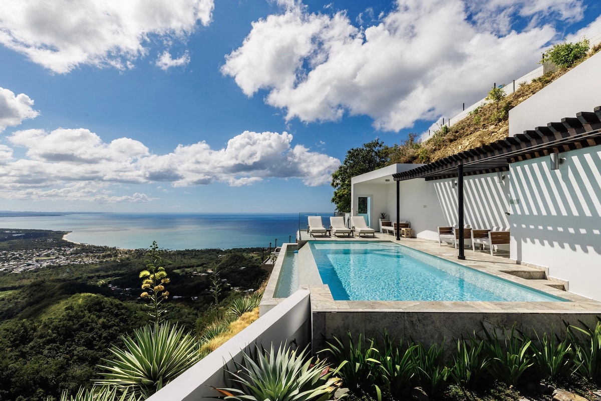 A serene infinity pool overlooks panoramic views of the coastline and rolling hills. The modern structure features a shaded lounge area with comfortable seating, surrounded by lush landscaping. The clear sky and scattered clouds enhance the tranquil atmosphere of the outdoor space.