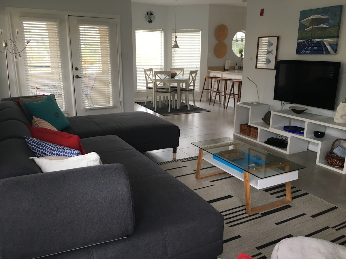 A modern living area features a dark sectional sofa adorned with colorful throw pillows. A glass coffee table sits on a patterned rug, while a flat-screen TV is mounted on a white shelf. Sunlight enters through sliding doors, illuminating a dining area in the background.