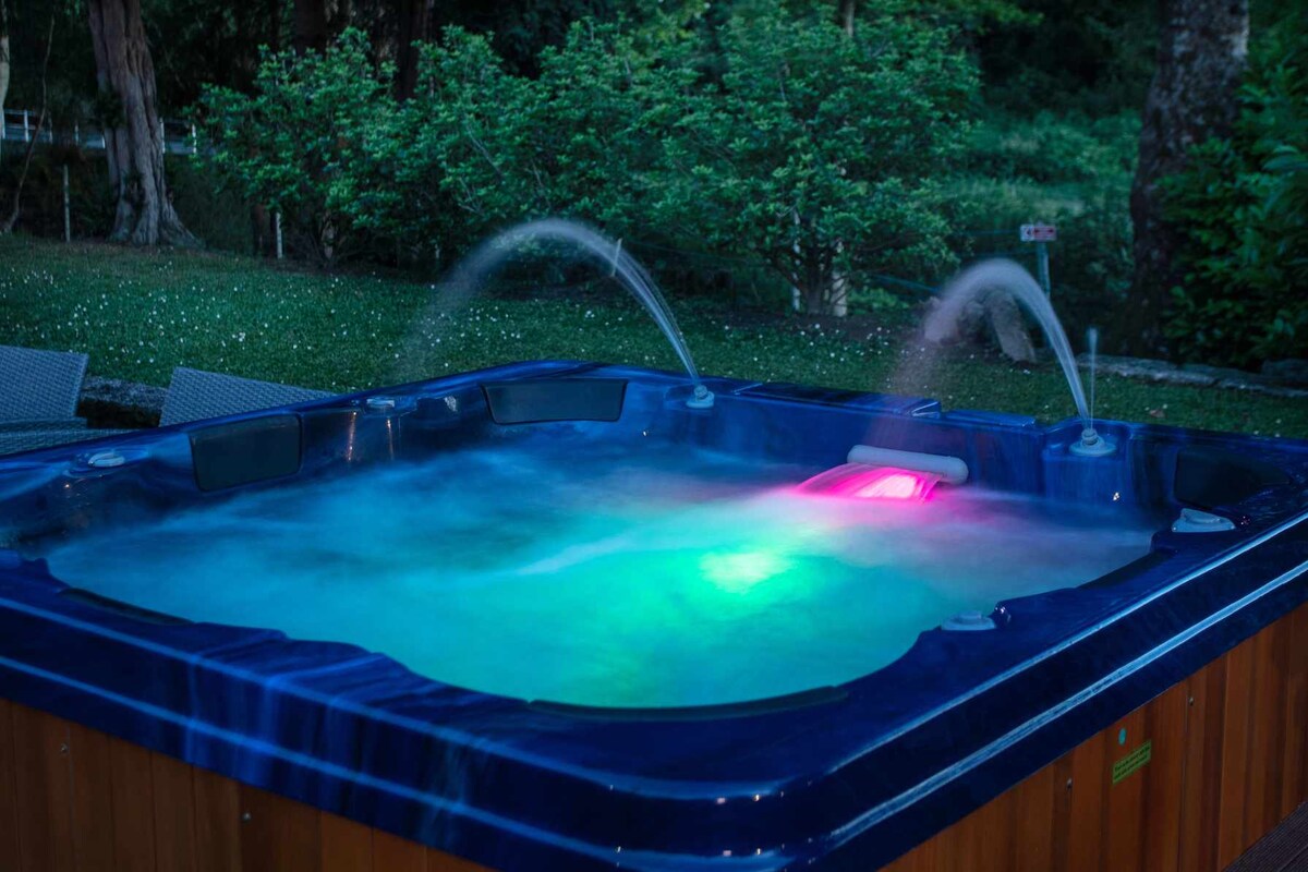 A hot tub is surrounded by greenery, featuring illuminated jets that create gentle water sprays. The tranquil water reflects soft blue and pink lights, providing a calming ambiance. Comfortable seating is nearby, enhancing the outdoor relaxation space.