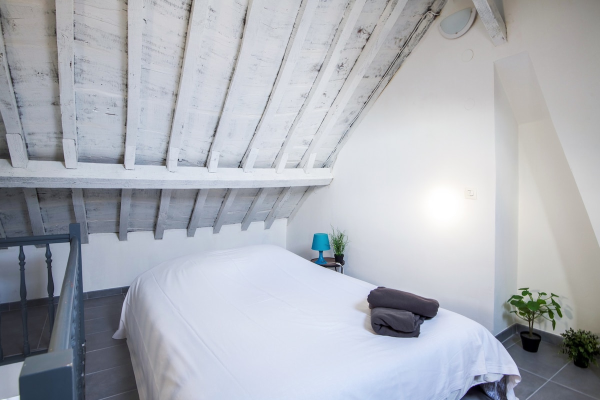 The sleeping area features a spacious bed with neatly arranged linens, complemented by two towels. The high, sloped ceiling showcases wooden beams, while a small blue lamp and potted plant add subtle touches of color to the bright, minimalist space.