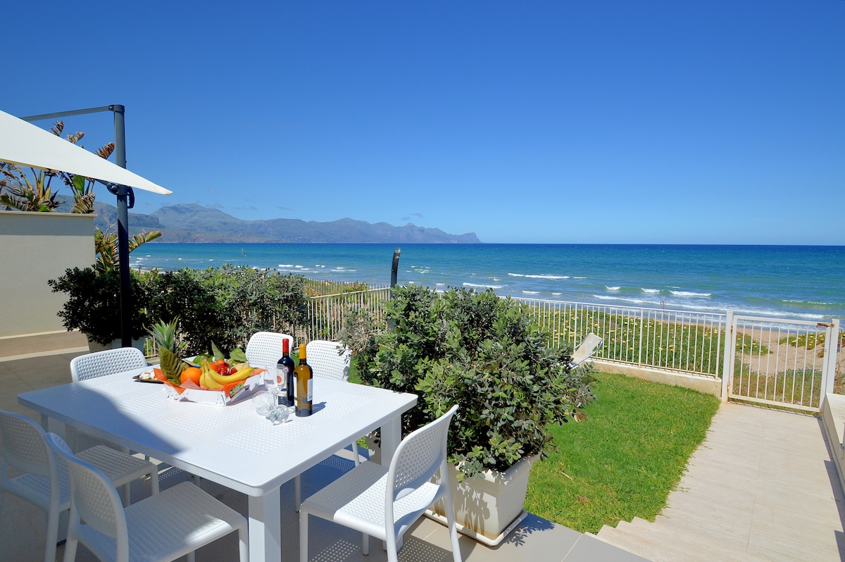 A dining area features a white table set with fresh fruit and drinks, surrounded by several white chairs. The space overlooks a serene sea view and lush greenery, with the ocean gently lapping against the shore under a clear blue sky.