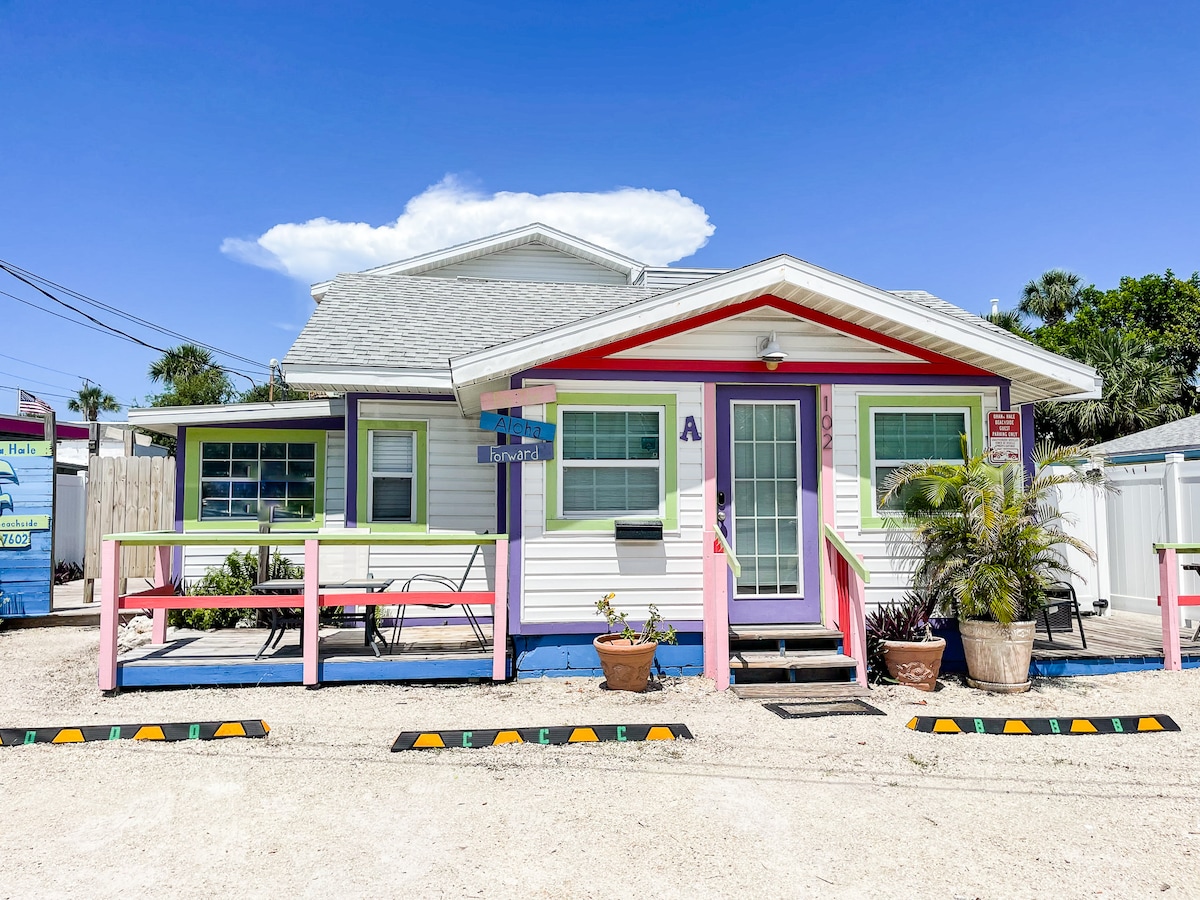 A charming single-story house features a colorful exterior with bright pink and purple accents. The entrance is framed by a small porch, which includes sitting areas and potted plants. The surroundings showcase a clear blue sky and a well-maintained pathway.
