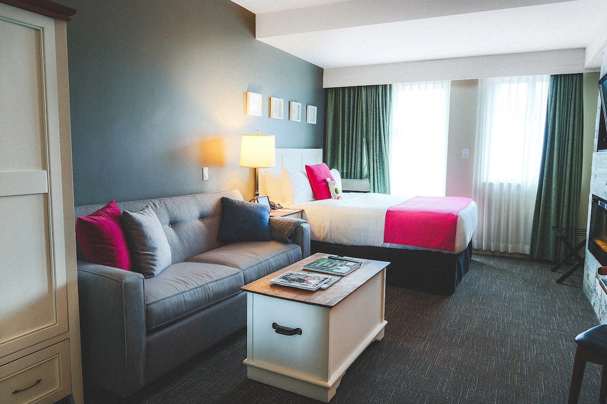 A bright room features a spacious King bed with crisp white linens and pink accents. A comfortable sofa is positioned nearby, complementing the playful decor. Natural light illuminates the space through large windows dressed with light green drapes, enhancing the room's cheerful atmosphere.