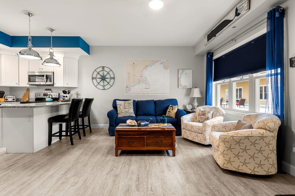 An open-concept living area features a navy blue sofa and two patterned chairs, enhancing relaxation. A wooden coffee table sits in the center. The kitchen area includes modern appliances and a bar counter with seating for four. Natural light filters in through a window dressed with blue curtains.