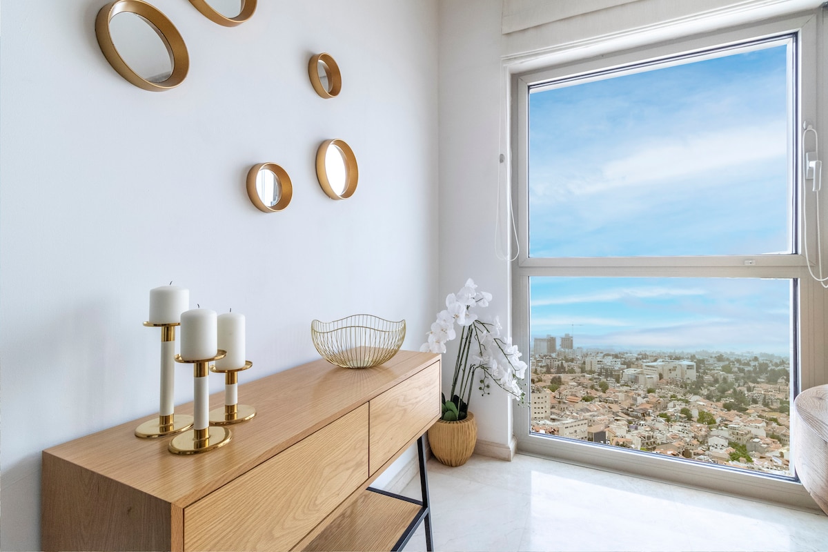 A wooden console table is positioned against a white wall, topped with elegant candles and a decorative bowl. Circular mirrors adorn the wall, reflecting light. A large window offers a panoramic view of the Jerusalem skyline, creating a sense of openness and connection to the surroundings.