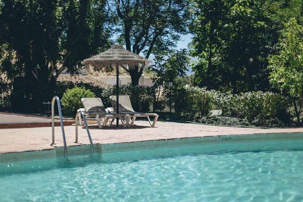 A clear swimming pool is framed by lush greenery and shaded by tall trees. Two lounge chairs are positioned beside the pool under a straw umbrella, offering a tranquil spot for relaxation.