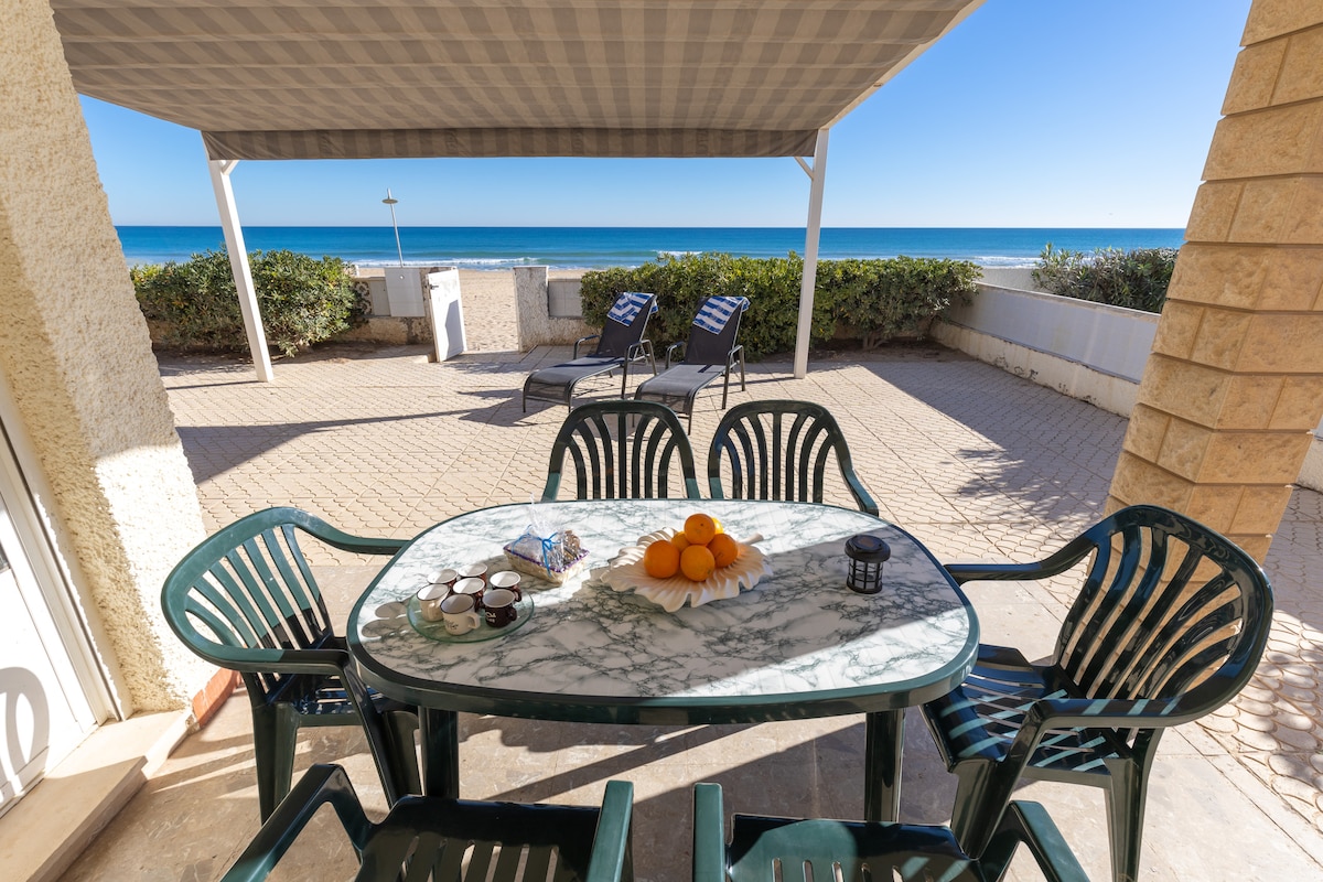An outdoor dining area is set on a spacious terrace, featuring a marble table surrounded by black and green chairs. A bowl of oranges sits in the center, with a view of the ocean visible beyond the terrace. Sun loungers are positioned to the side.