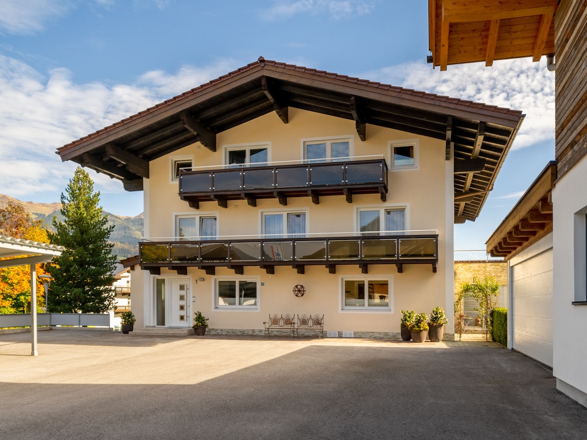 The exterior of the apartment house features a three-story building with a sloped roof. Multiple balconies adorned with wooden railings provide outdoor space. Fresh greenery surrounds the entrance, and a designated parking area is visible in the foreground.