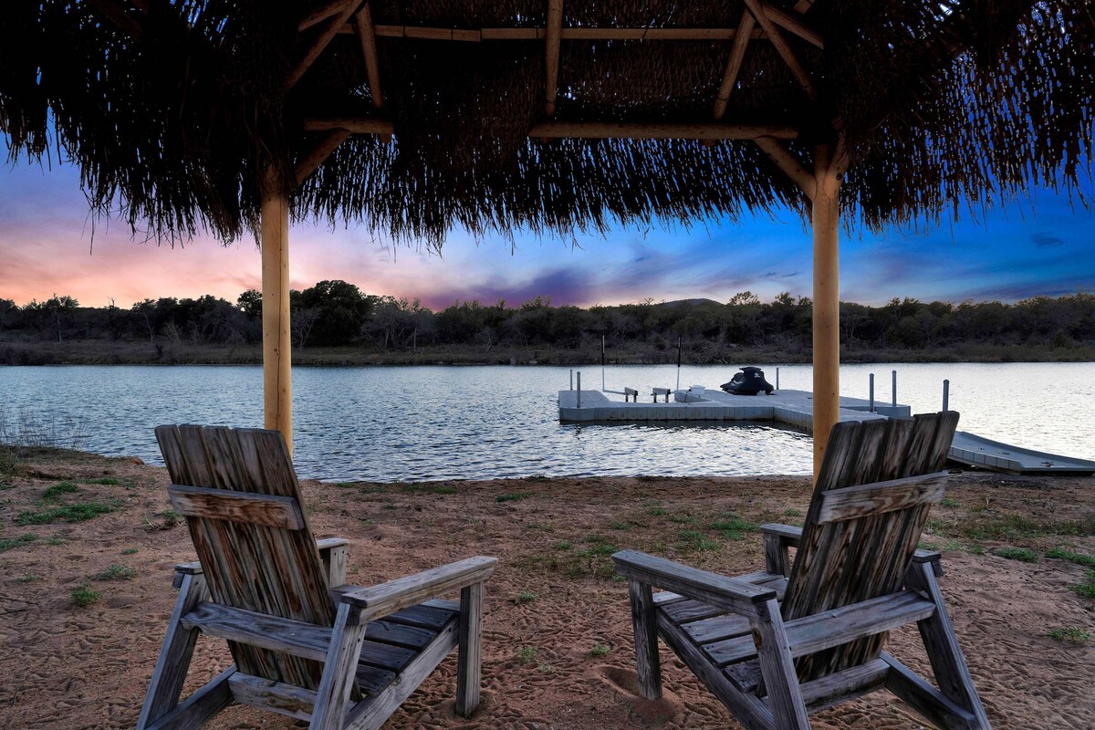 A covered pergola provides shade over two rustic wooden chairs, positioned on a sandy beach. In the background, a floating dock is visible on the calm water, with the colorful sky reflecting above during twilight hours.