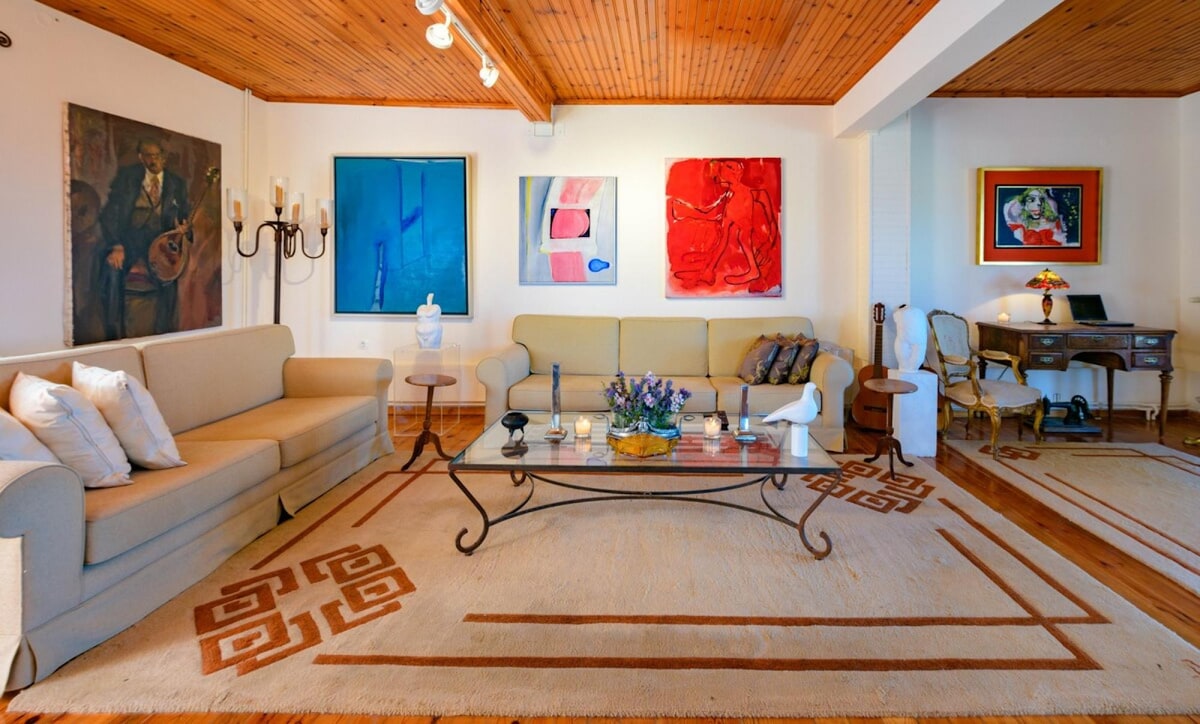 A spacious living area features a light-colored sofa and elegant armchairs surrounding a glass coffee table. Colorful artworks adorn the walls, while a wooden desk with an ornate chair complements the room's ambiance. A patterned rug adds warmth to the hardwood floor.