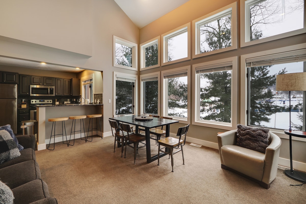 A spacious living area is highlighted by large windows that allow natural light to fill the space. A dining table is positioned in the center, surrounded by chairs. A modern kitchen is visible in the background, offering a welcoming atmosphere. A cozy couch and armchair complement the room.
