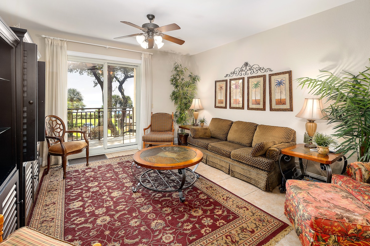 A spacious living area features a patterned rug and a coffee table at the center. Large glass doors lead to a private balcony, allowing natural light to fill the room. Comfortable seating includes a sofa, armchair, and a patterned chair, complemented by decorative plants.